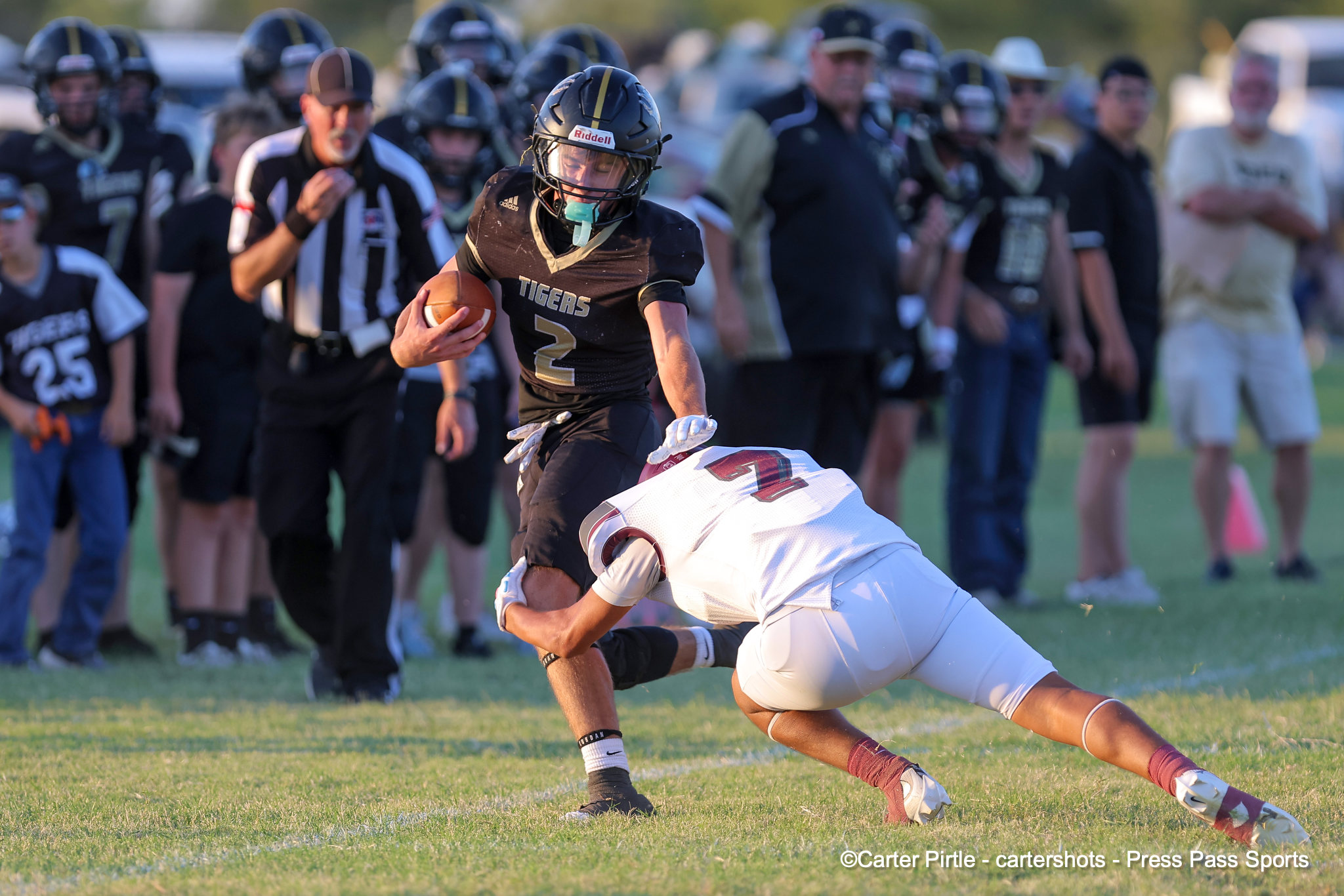 Football player in a black Tigers jersey (number 2) runs with the ball while a white-uniform defender dives to tackle, with a crowd in the background on a grassy field.
