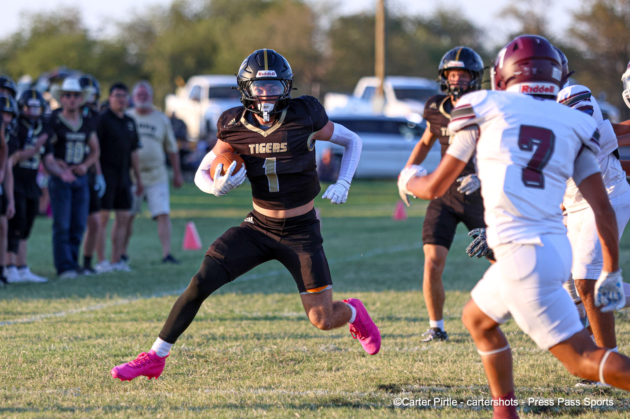 Football player in a black Tigers jersey runs with the ball during a game on a grassy field, white-uniformed players giving chase in the background.