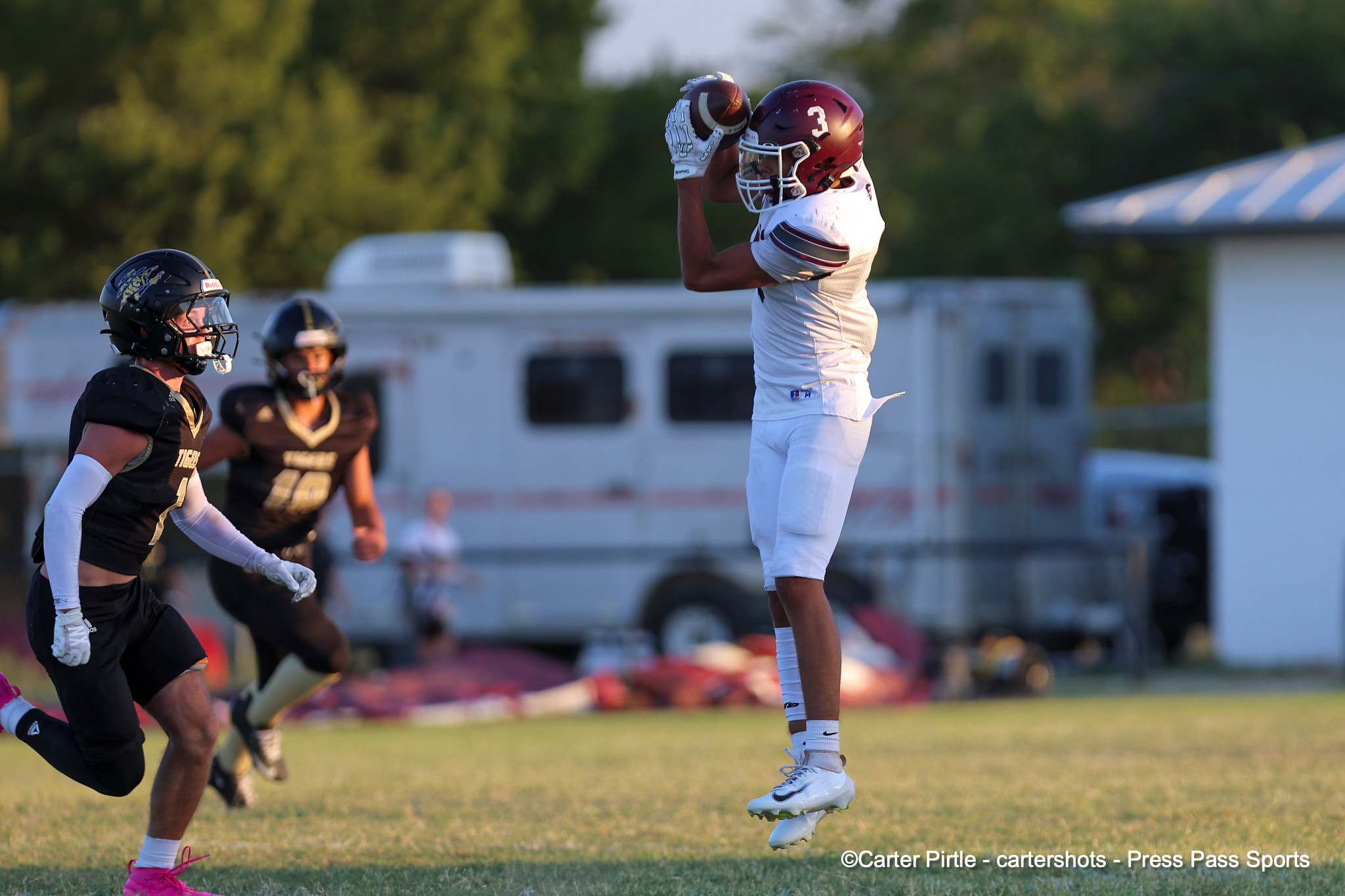 Football player in a white uniform leaps to catch a pass as black-clad defenders move toward him on a sunny field.