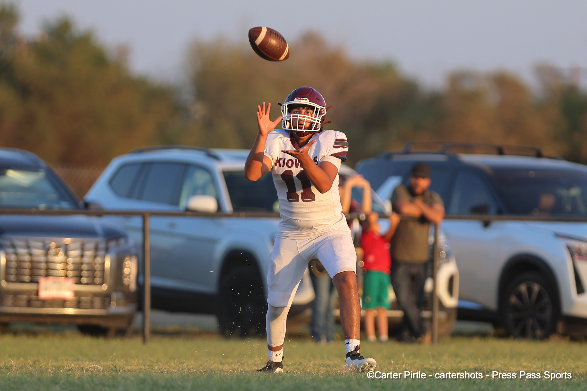 Football player in a white uniform and maroon helmet reaches up to catch a pass midair on a sunny field.