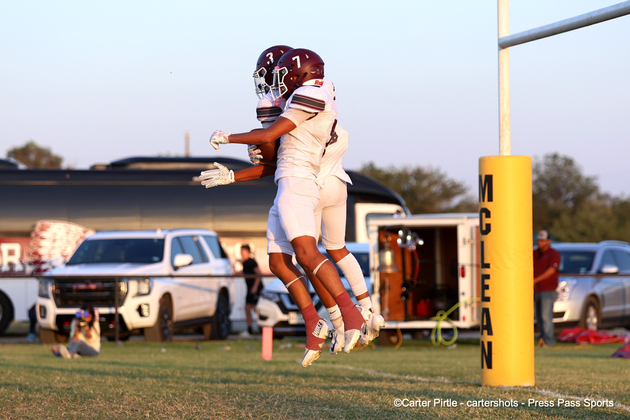 Two football players in white uniforms jump together near the end zone goalpost, helmets maroon, celebrating on a sunny field.