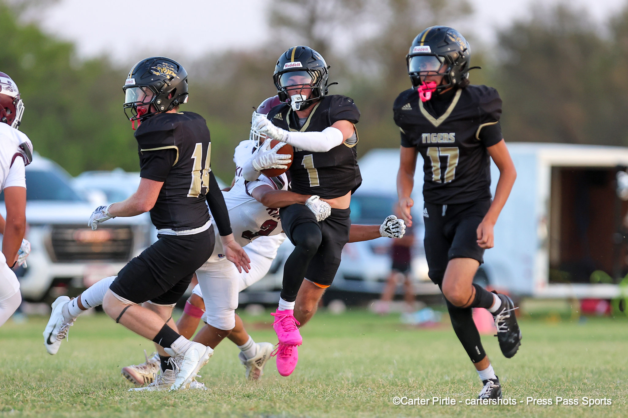 Group of football players in black uniforms chase the ball on a grass field; center player clutches the ball while teammates defend.