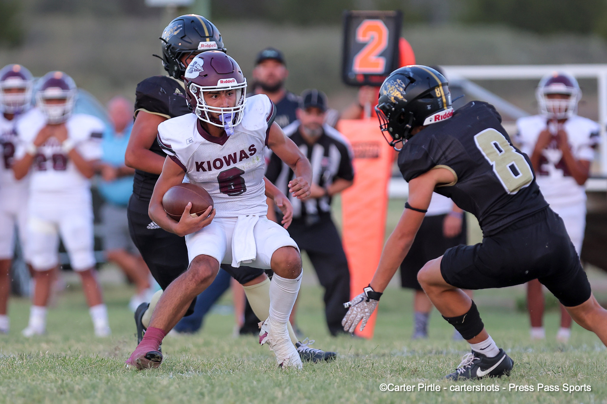 Young football player in a white Kiowas jersey runs with the ball while defenders close in on a grassy field.