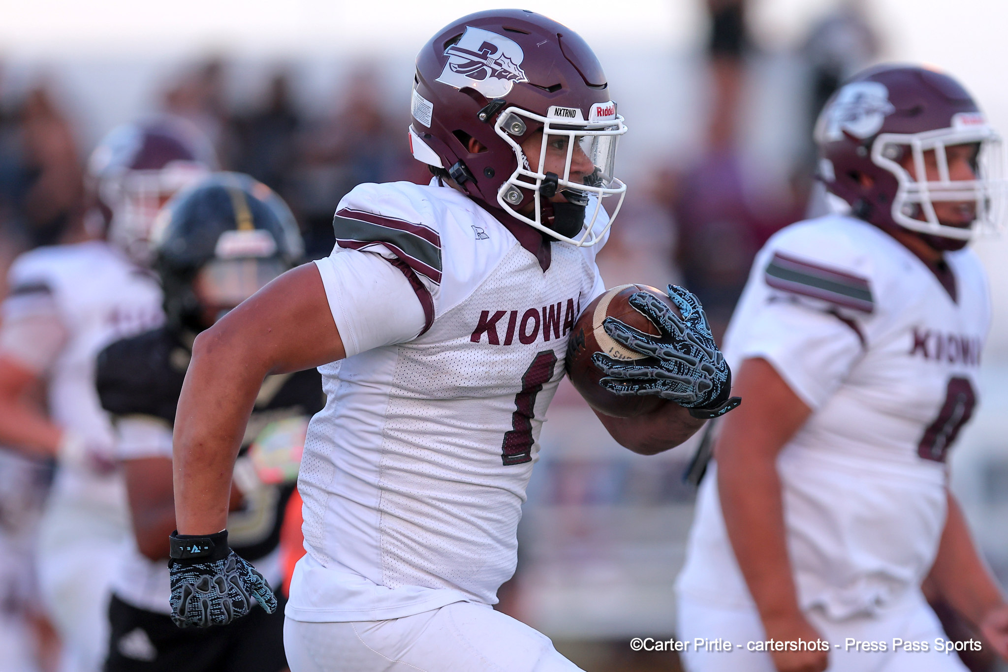 Football player in white Kiowa uniform running with the ball, maroon helmet and patterned gloves, action shot on the field with teammates blurred in the background