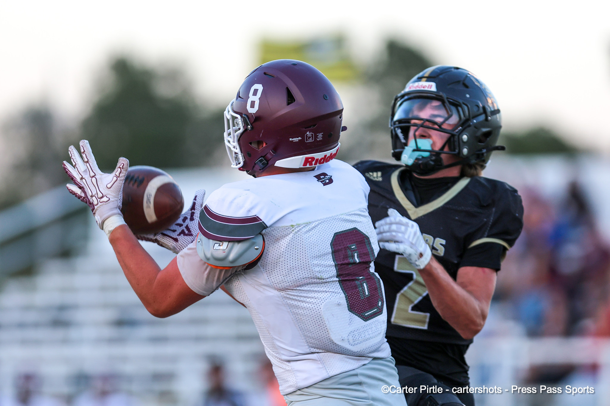Two American football players collide as one in a white jersey with a maroon helmet reaches for the ball while a defender in dark gear pursues.
