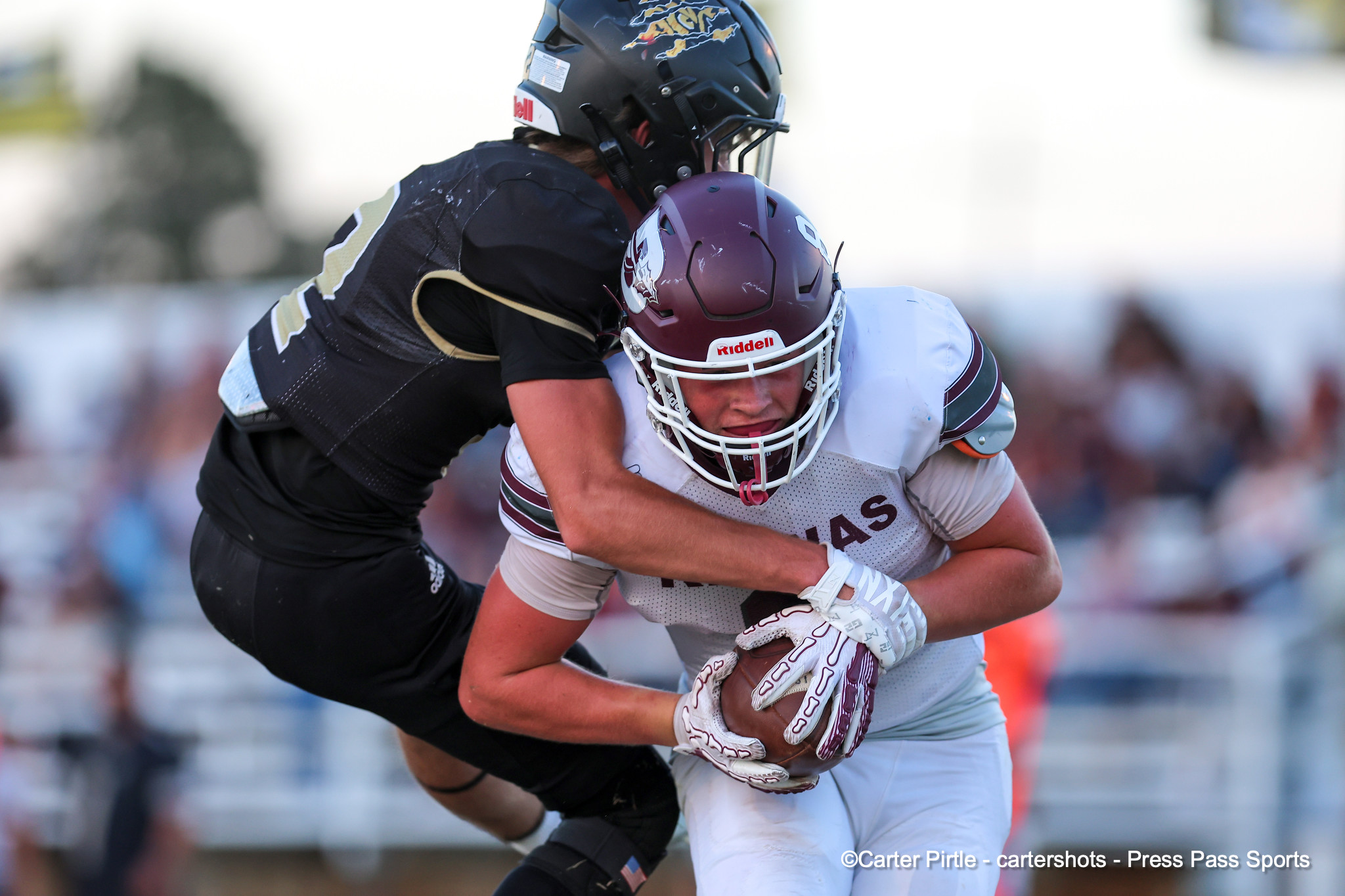 Two football players collide: a defender in maroon helmet and white jersey tackles a black-clad opponent near the chest on a sunny field