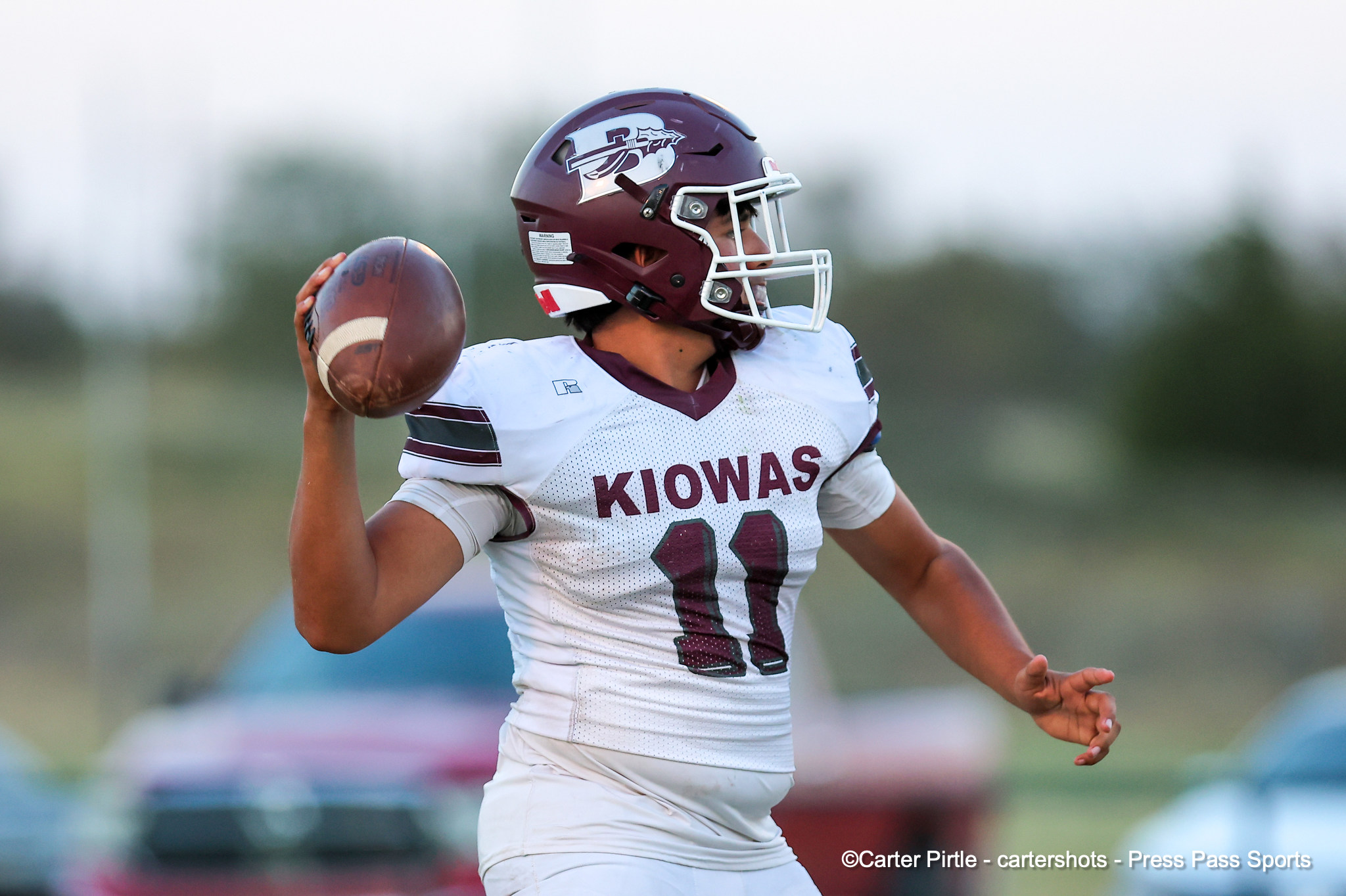 Quarterback in a maroon helmet and white Kiowas uniform throws a football on the field.