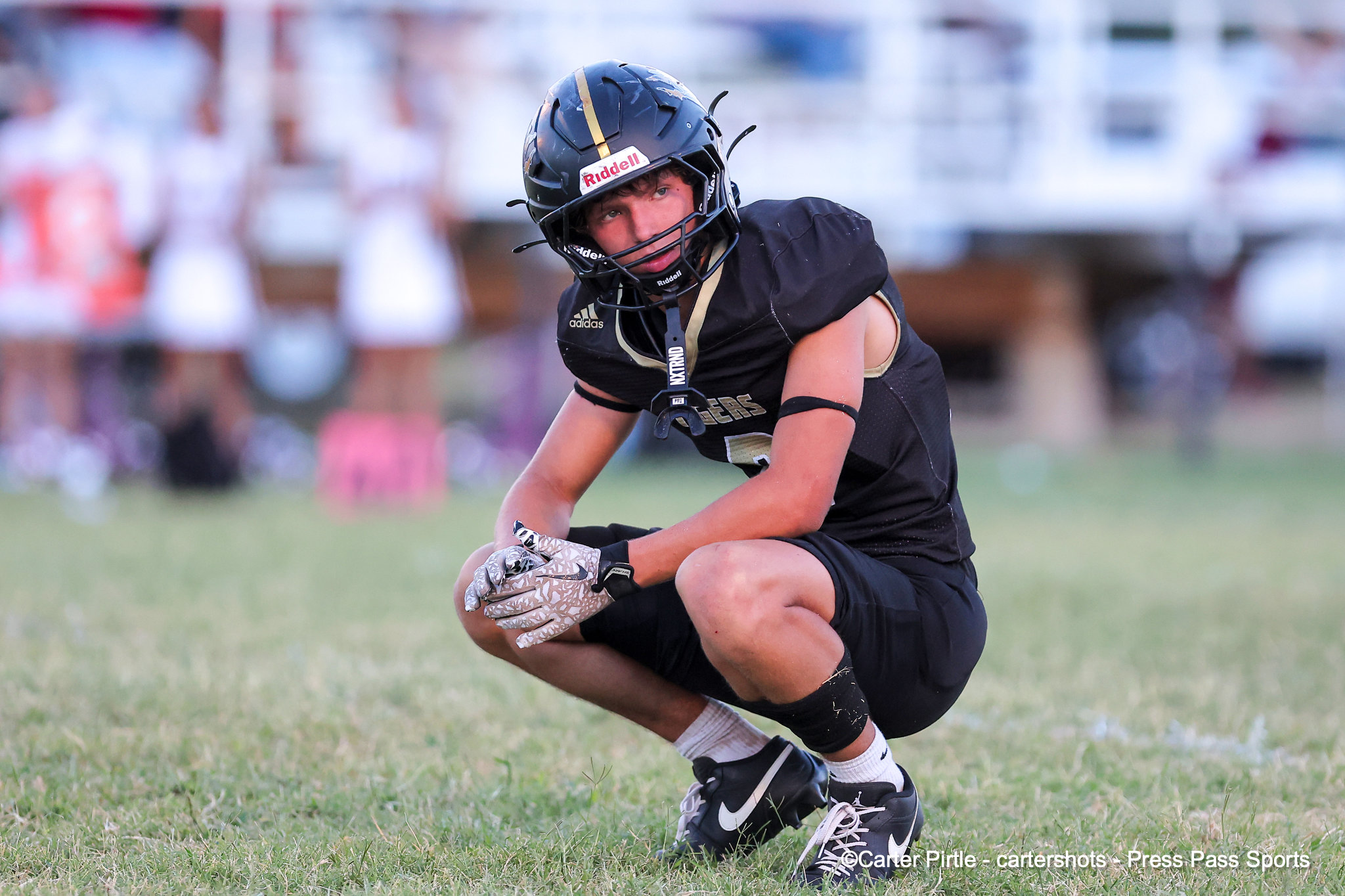 Football player in a black uniform crouches on a grassy field, wearing a helmet and gloves, ready for a snap.