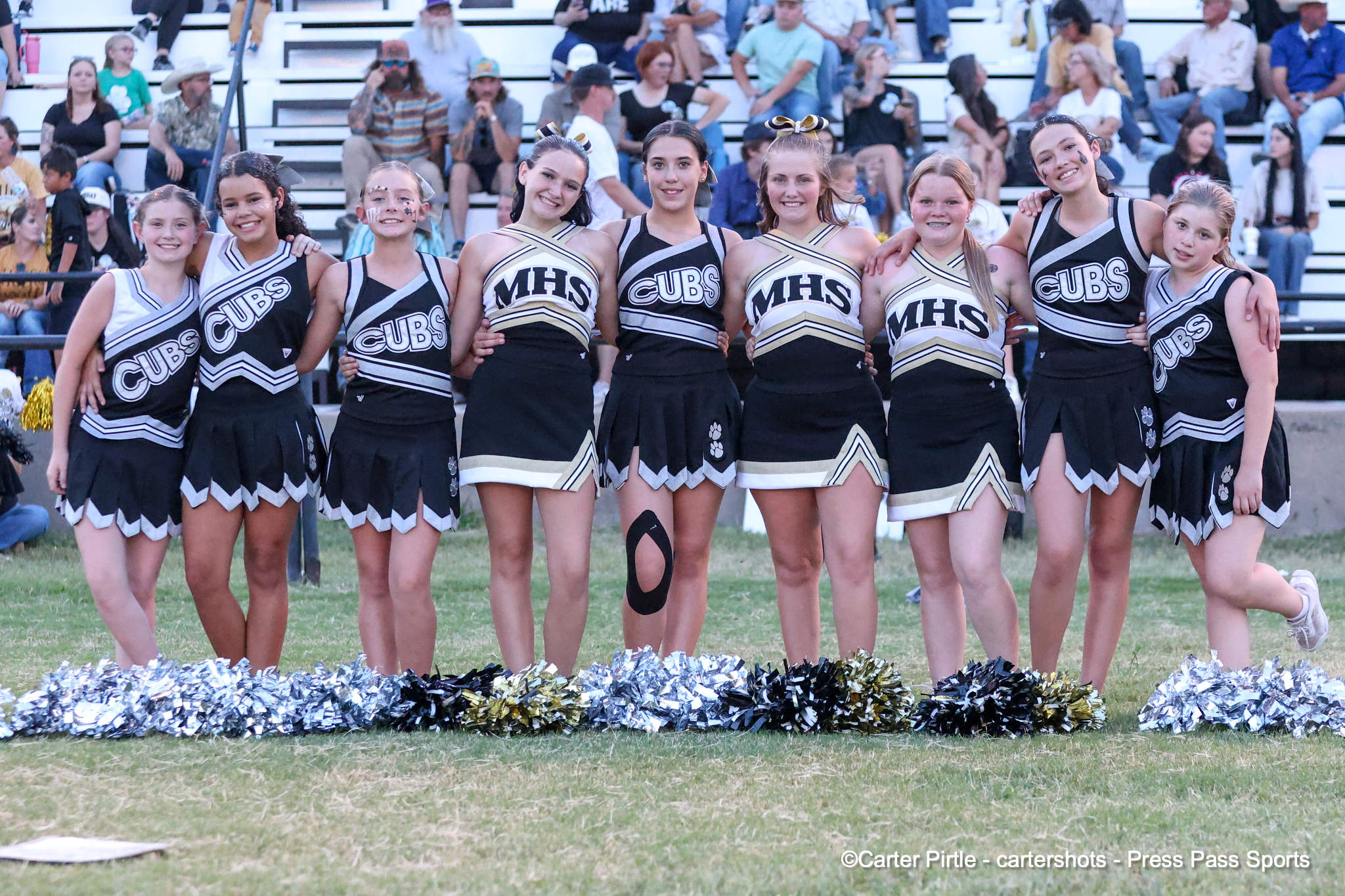 Group of female cheerleaders in black-and-gold uniforms stands with arms around each other on a field, bleachers full of spectators behind them.