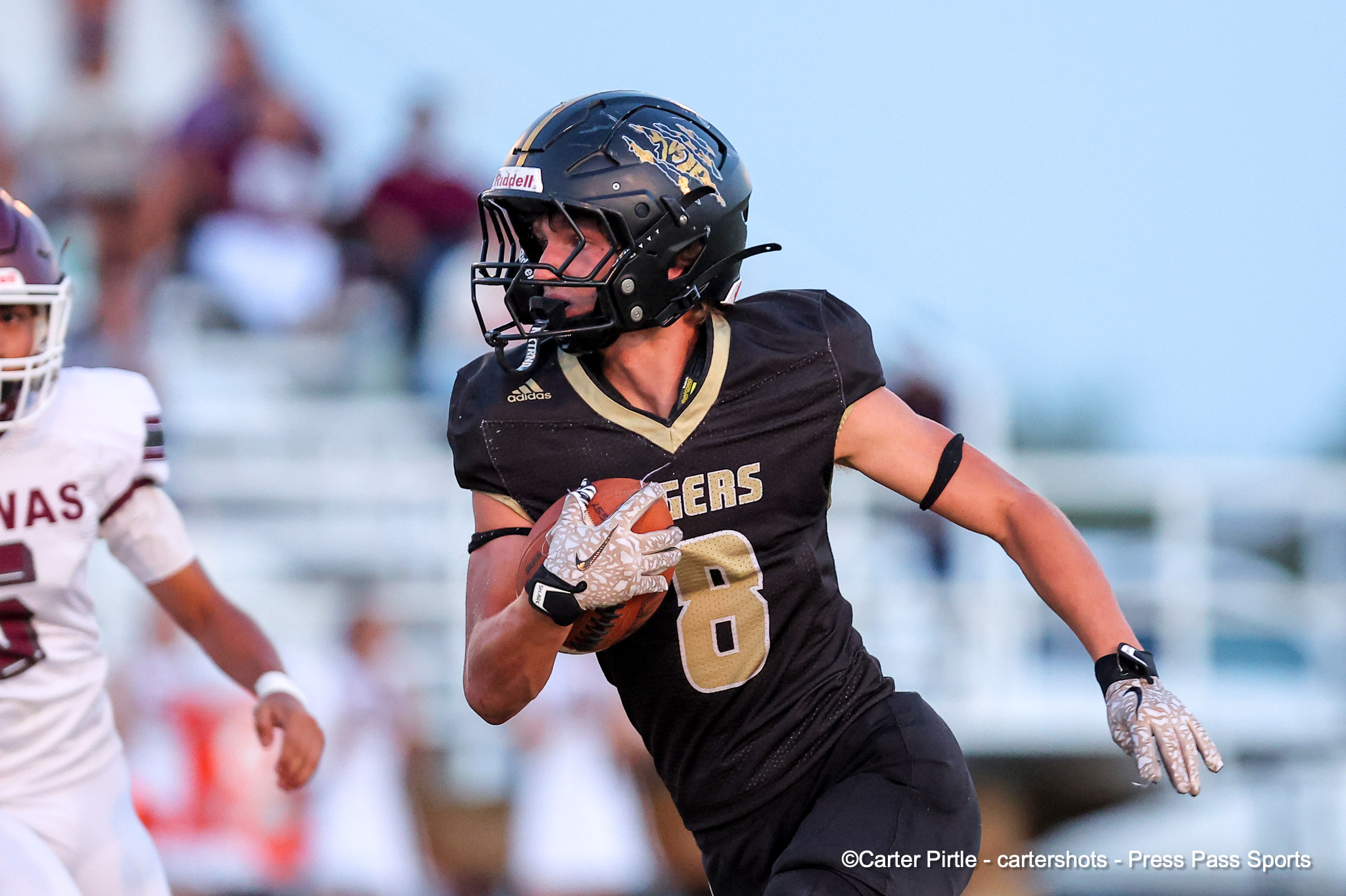 Action shot of a football player in a black and gold uniform running with the ball during a game, helmet on, with a blurred crowd in the background.