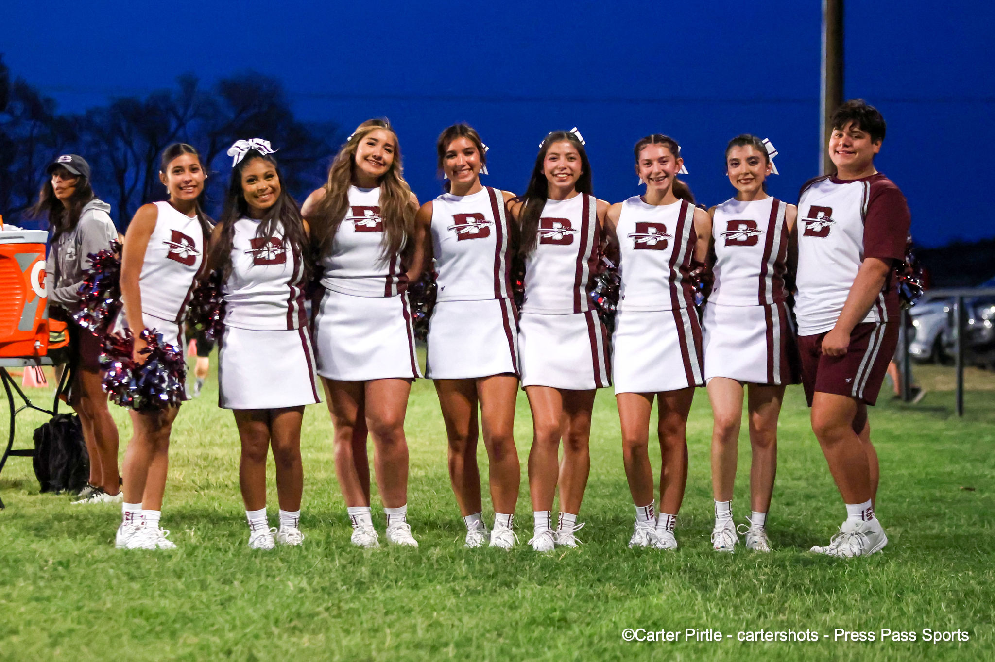 Cheerleading squad posing together on a grassy field at dusk in white and maroon uniforms with bows.