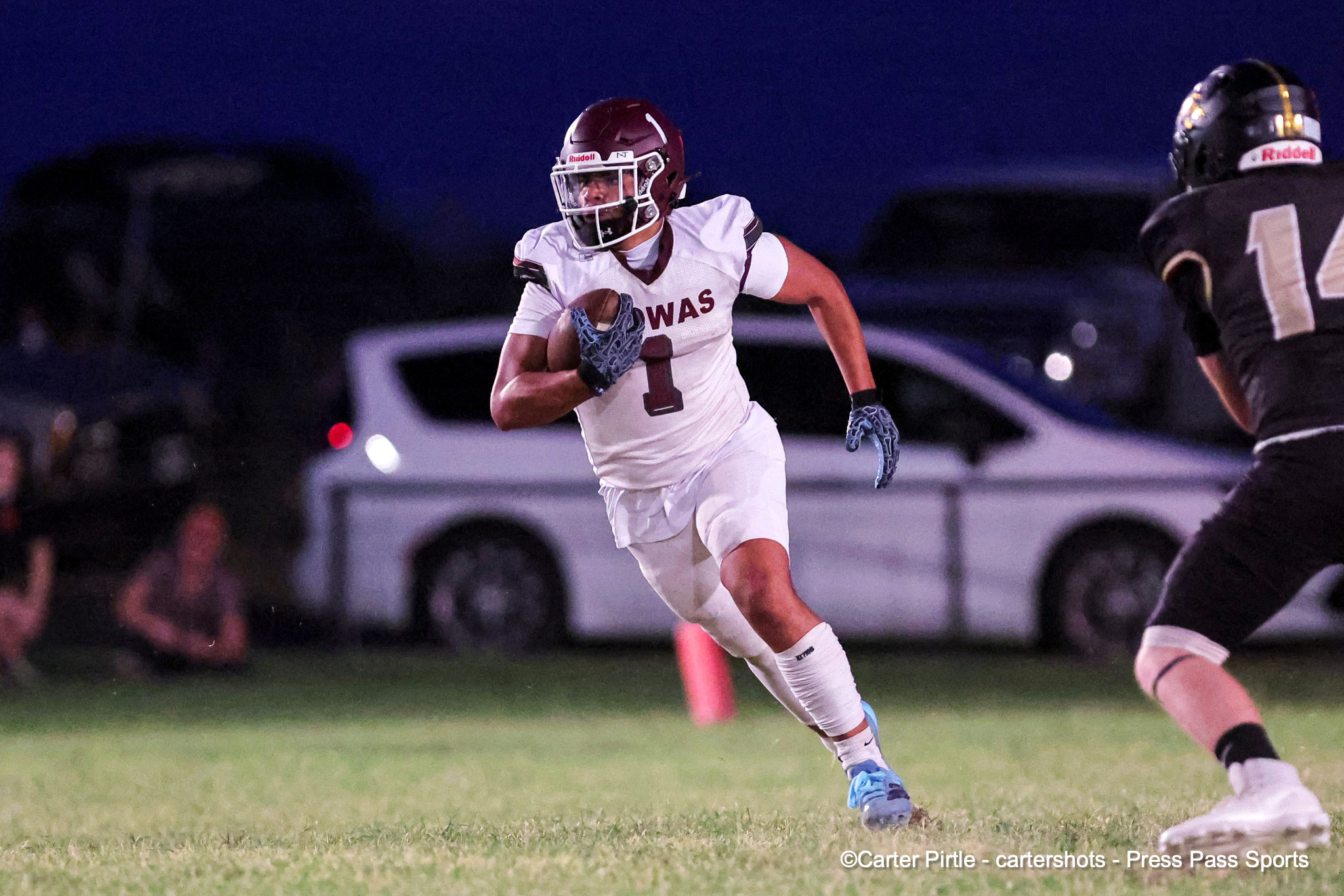 Football player in white jersey with maroon number 1 carries the ball while running on a night game, pursued by a defender in black.