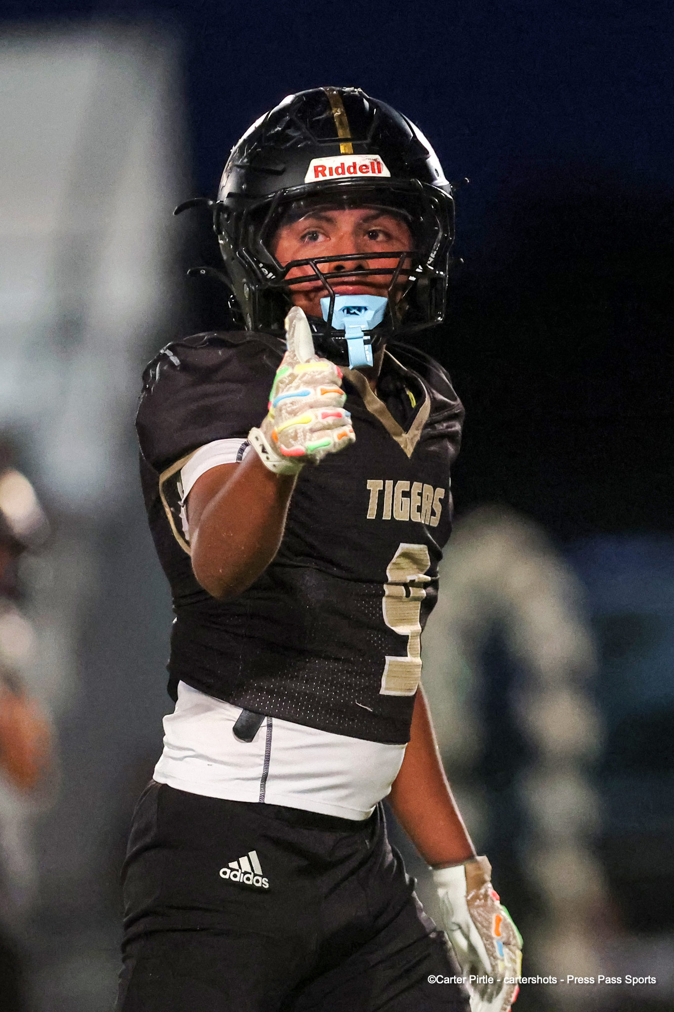 Football player in a black Tigers jersey gives a thumbs-up at night, wearing a Riddell helmet and colorful gloves.