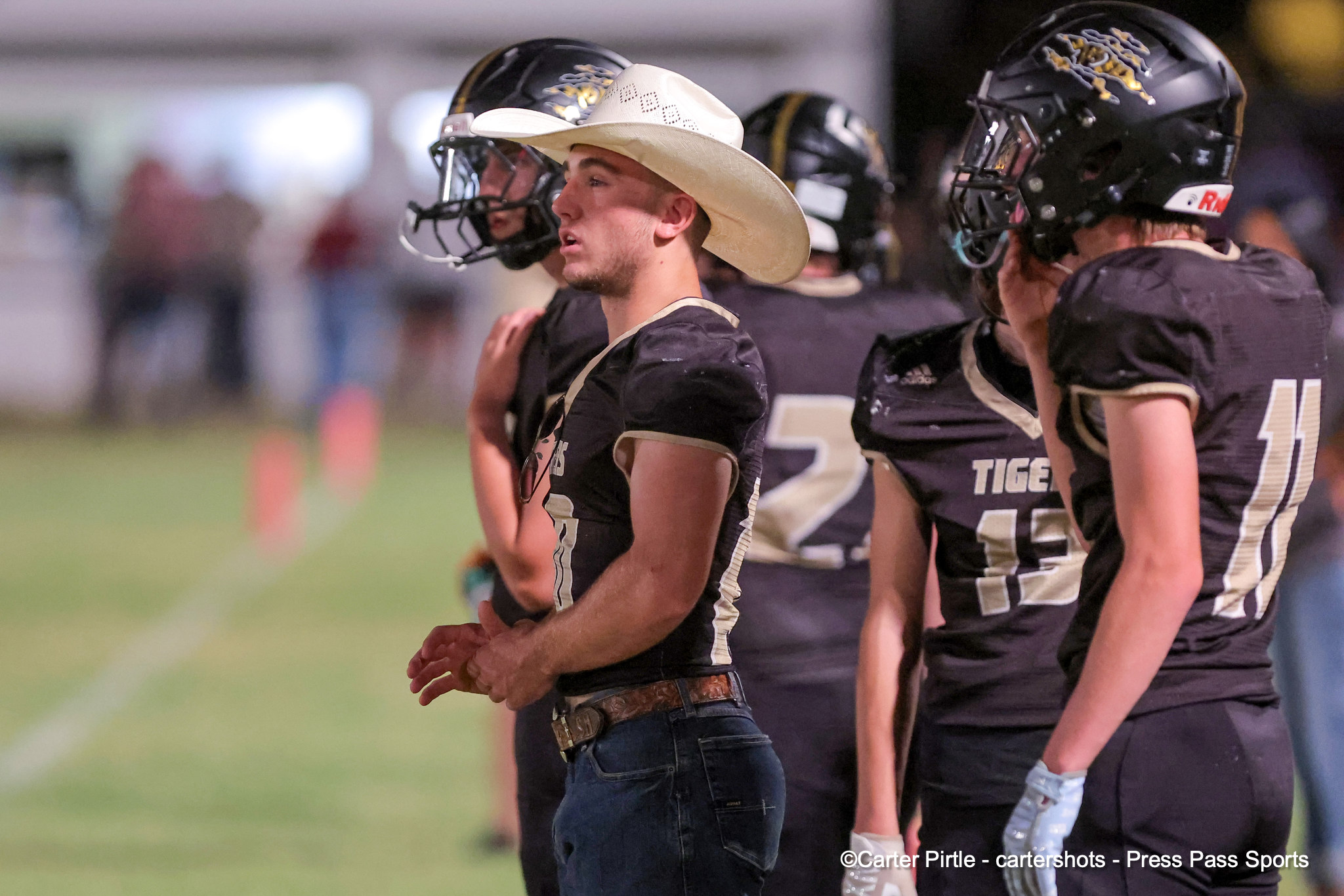 Football players in black uniforms stand together on the sideline; central figure wears a white cowboy hat and looks to the left.