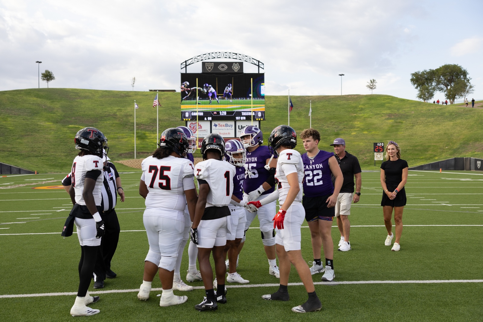 THS-CHS-FB-4 | Press Pass Sports High school football players from white and purple teams huddle on the field before the game, with a large scoreboard and grassy hills in the background.