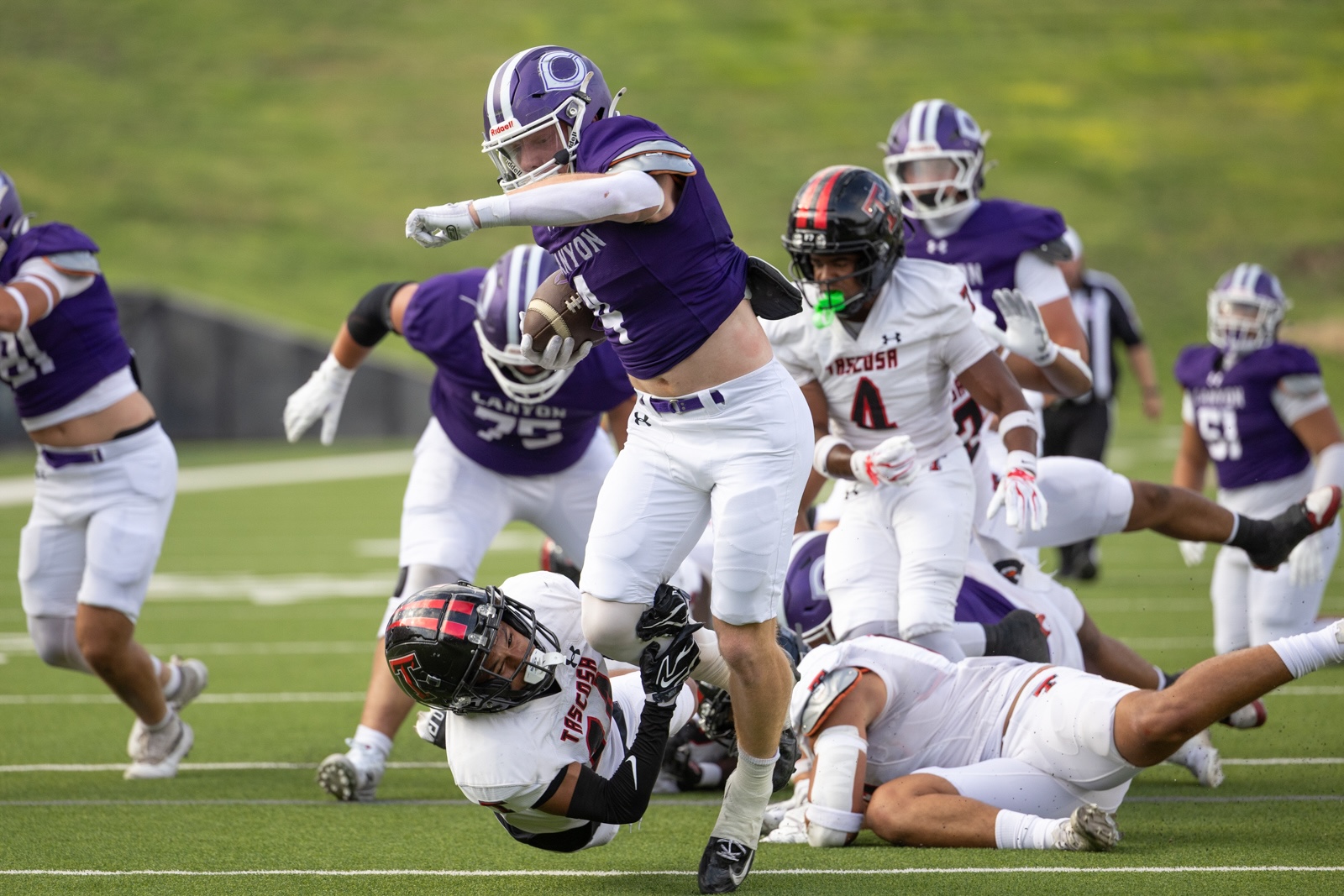 THS-CHS-FB-8 | Press Pass Sports A football player in a purple jersey leaps over a tackle while holding the ball during a game, with white-jersey defenders and teammates around on a green field.
