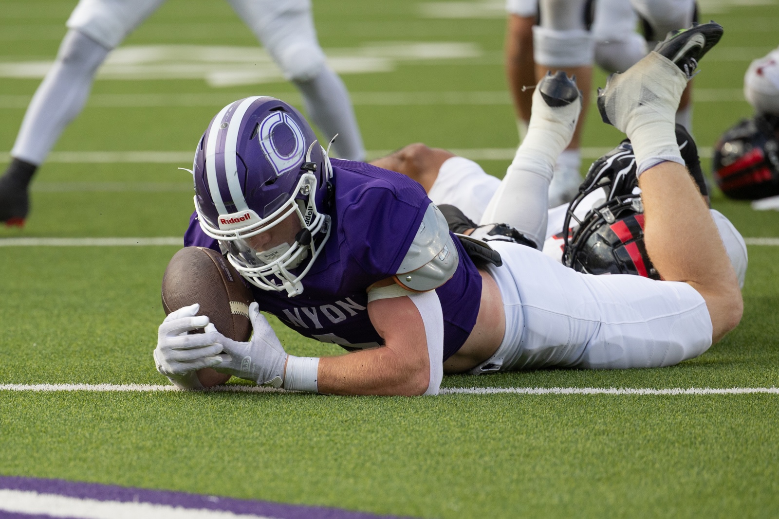 THS-CHS-FB-9 | Press Pass Sports Purple-uniform football player lunges and clutches a football while lying on a green field, white gloves gripping the ball as teammates scramble nearby.