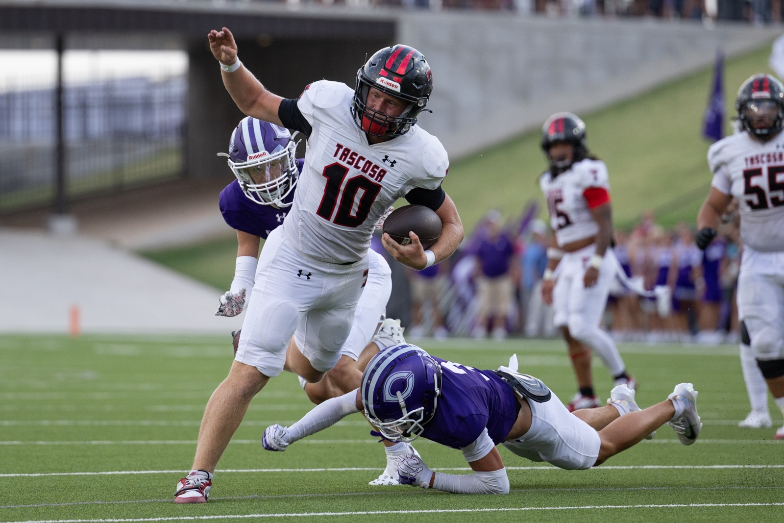 THS-CHS-FB-14 | Press Pass Sports TASCOSA running back #10 carries the football past a diving purple defender on a green football field, with teammates and spectators in the background.