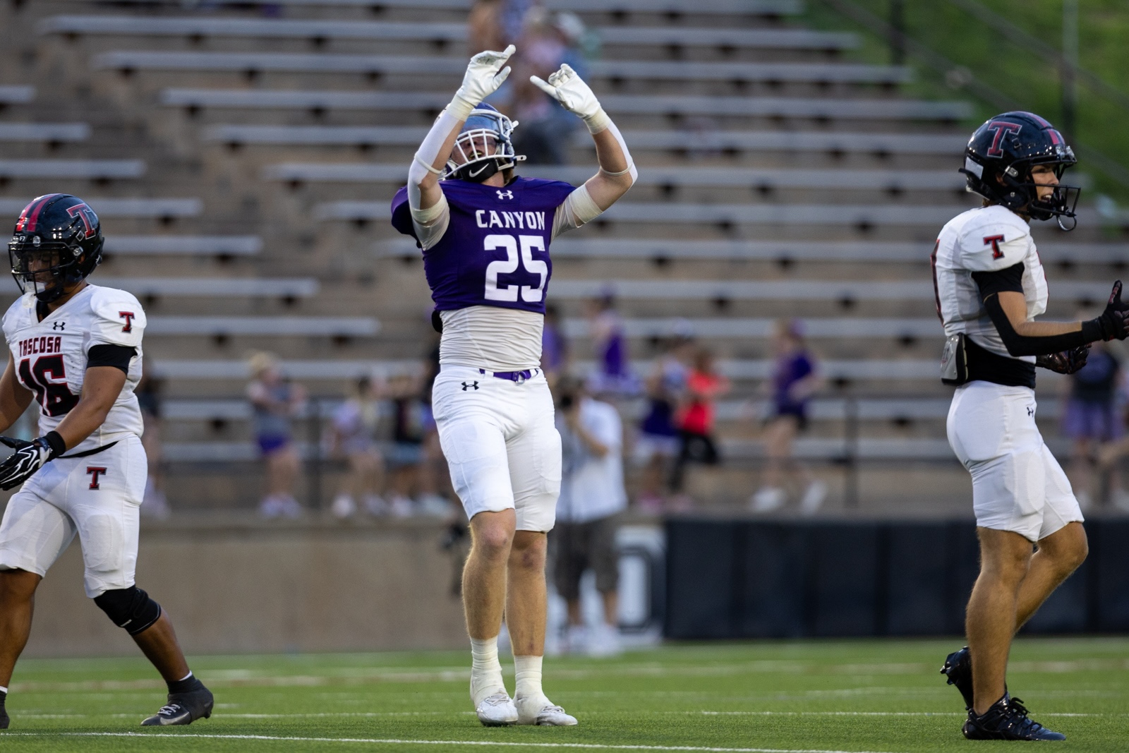 THS-CHS-FB-30 | Press Pass Sports Football player in purple Canyon jersey #25 raises his arms in celebration on the field, flanked by teammates in white uniforms with red logos, bleachers in the background.