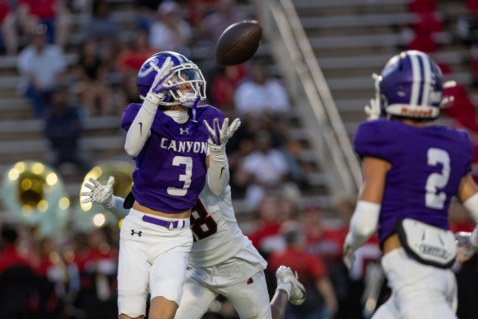 THS-CHS-FB-33 | Press Pass Sports Two Canyon football players in purple jerseys reach for a high football in midair during a game; #3 on the left, crowd visible in the stands behind them.