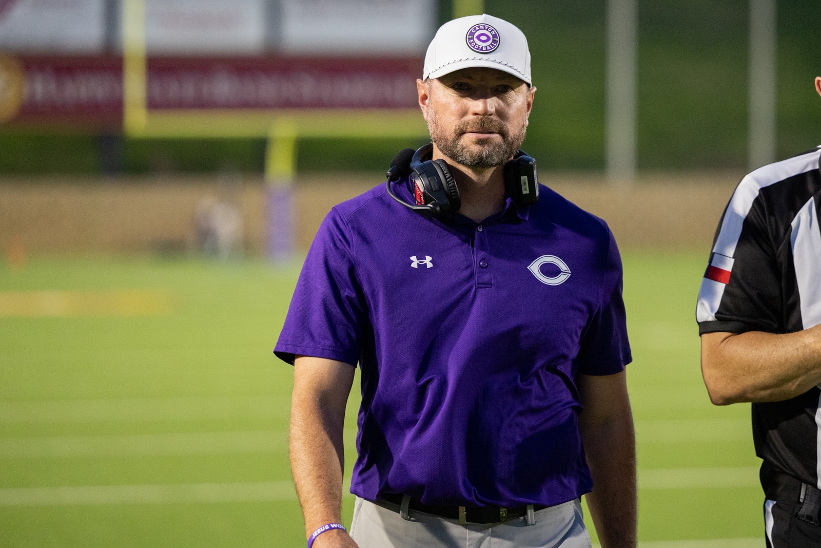 THS-CHS-FB-35 | Press Pass Sports Coach wearing a purple polo with headset around his neck on a football field, white cap, focused expression.