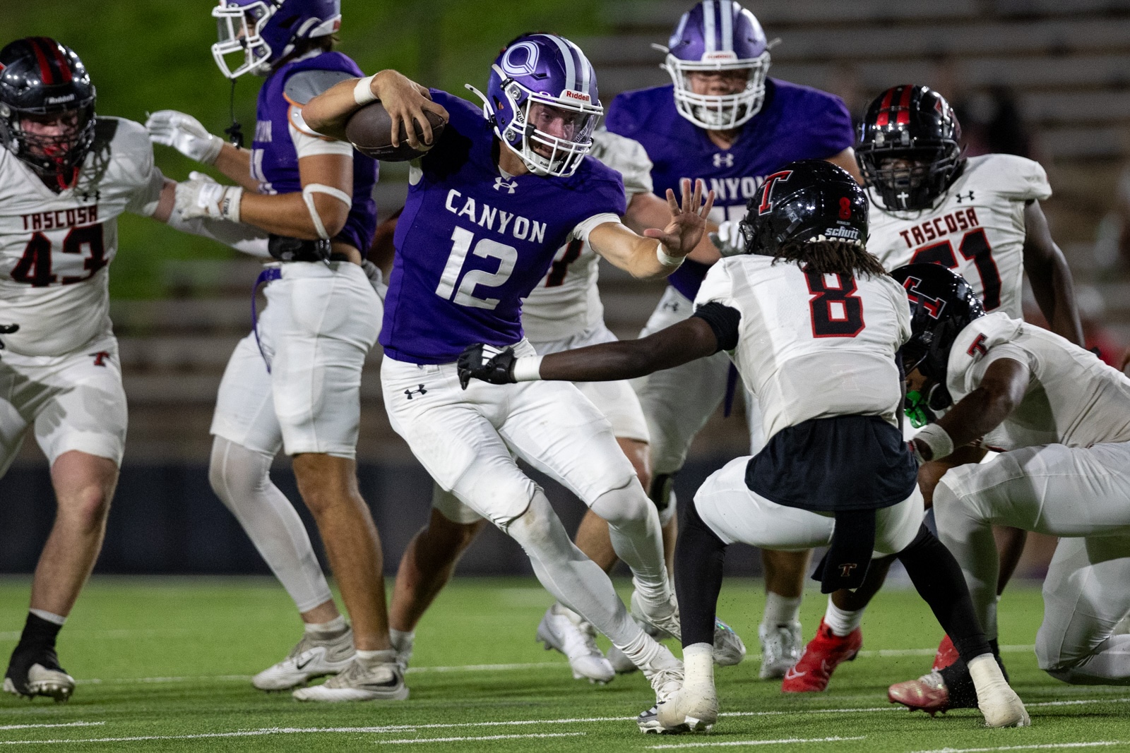 THS-CHS-FB-40 | Press Pass Sports Canyon quarterback #12 in purple, holding the ball and moving forward as defenders close in on a night game on a grassy field.