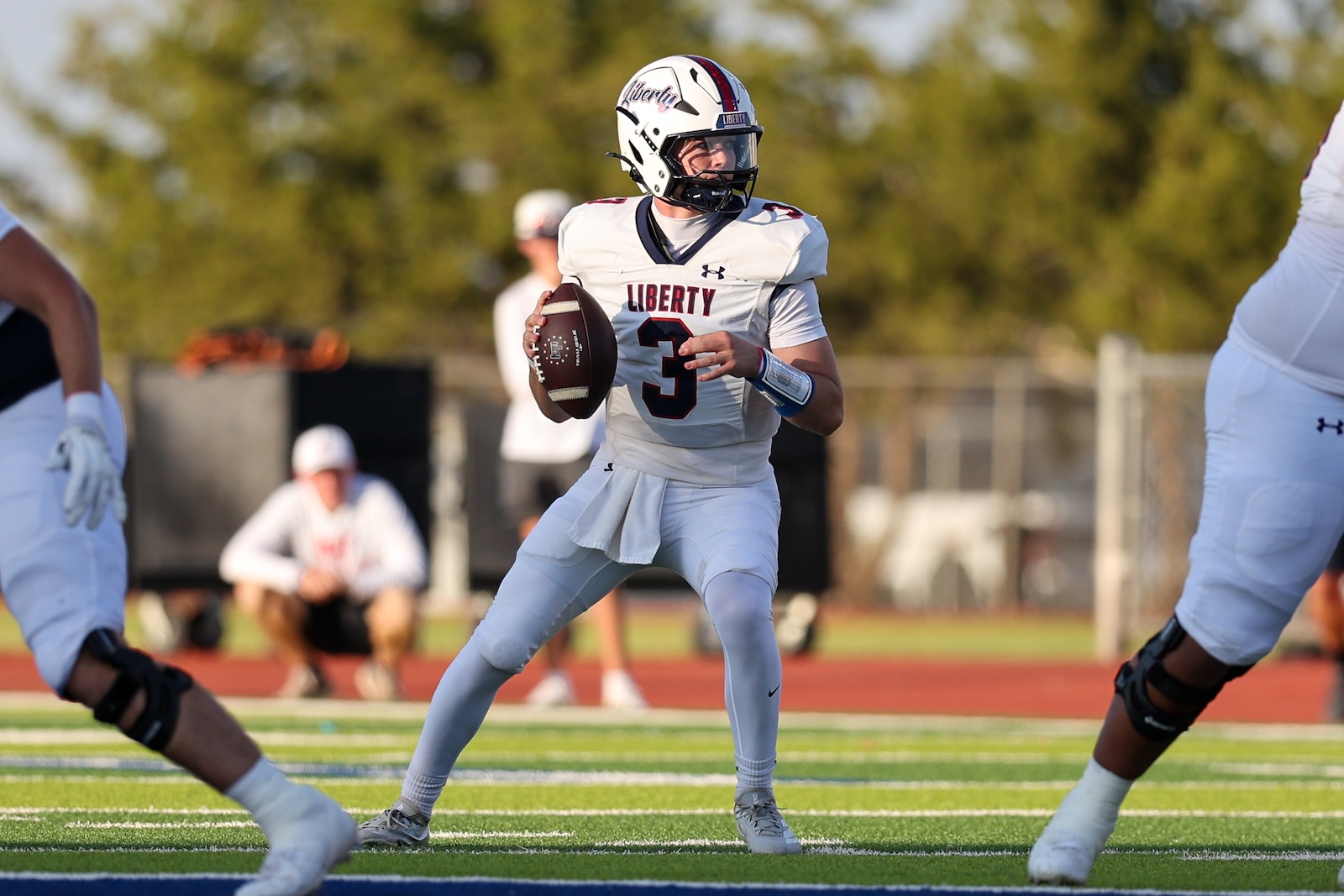 Seabolt Chase-16418 | Press Pass Sports Quarterback in white Liberty uniform (#3) grips a football and looks for an opening during a game on a sunny field, with teammates and a sideline in the background.