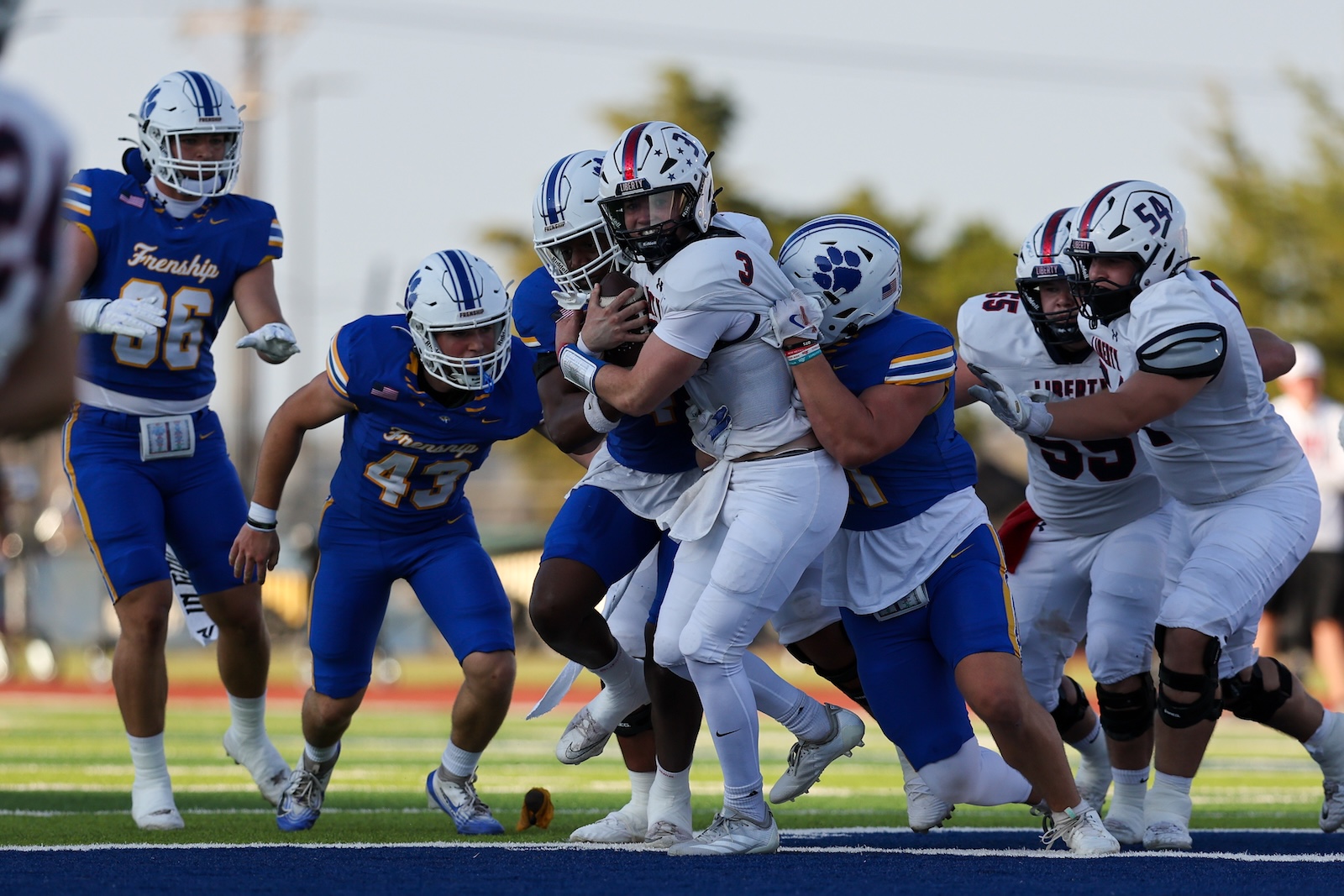 Seabolt Chase-16539 | Press Pass Sports Football play in progress: a white-uniform ball carrier is tackled by multiple blue-uniform defenders while teammates block nearby on a green turf field.