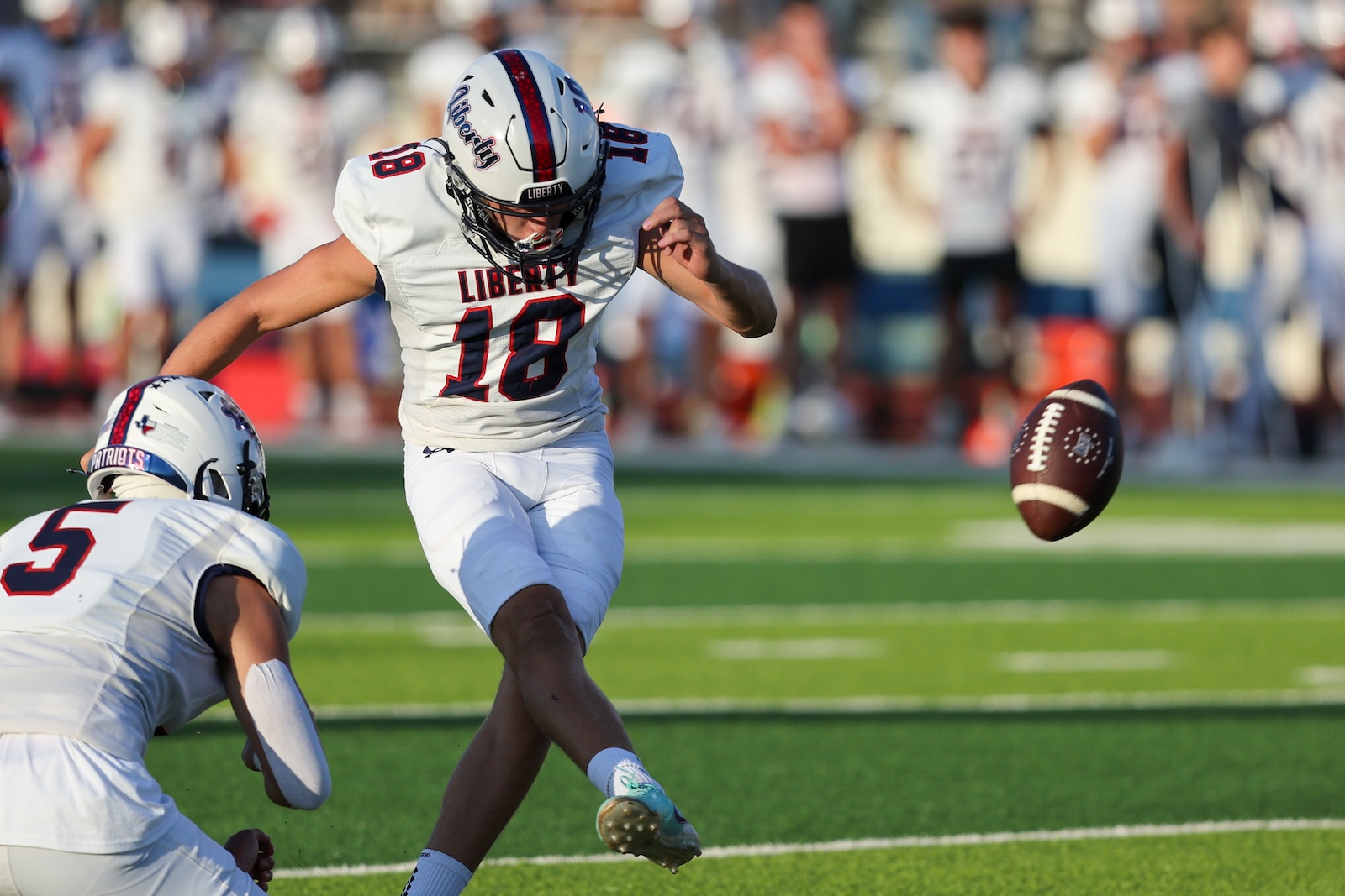 Seabolt Chase-16699 | Press Pass Sports Liberty player #18 in white uniform leaps toward an incoming football on a green field as a teammate looks on in the foreground.