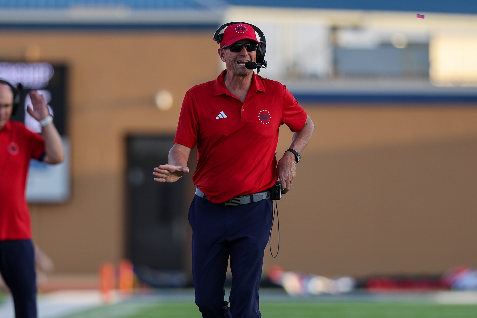 Seabolt Chase-17071 | Press Pass Sports Male coach in a red polo shirt and cap with headset speaking into a microphone on a football field during practice/game.