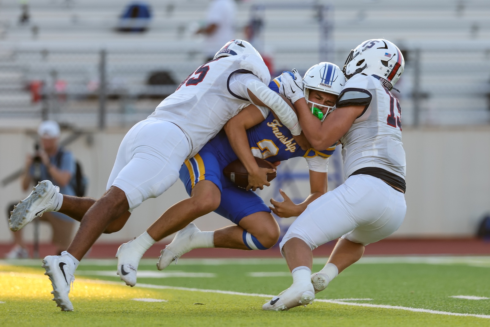 Seabolt Chase-17110 | Press Pass Sports American football scene: a ball carrier in blue and gold is tackled by two white-uniform players in a crowded, high-contact moment on the field.