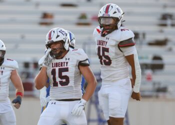Seabolt Chase-17193 | Press Pass Sports Liberty football players in white uniforms on the field; foreground player #15 adjusting his helmet with another teammate nearby.