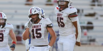 Liberty football players in white uniforms on the field; foreground player #15 adjusting his helmet with another teammate nearby.