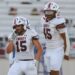 Seabolt Chase-17193 | Press Pass Sports Liberty football players in white uniforms on the field; foreground player #15 adjusting his helmet with another teammate nearby.