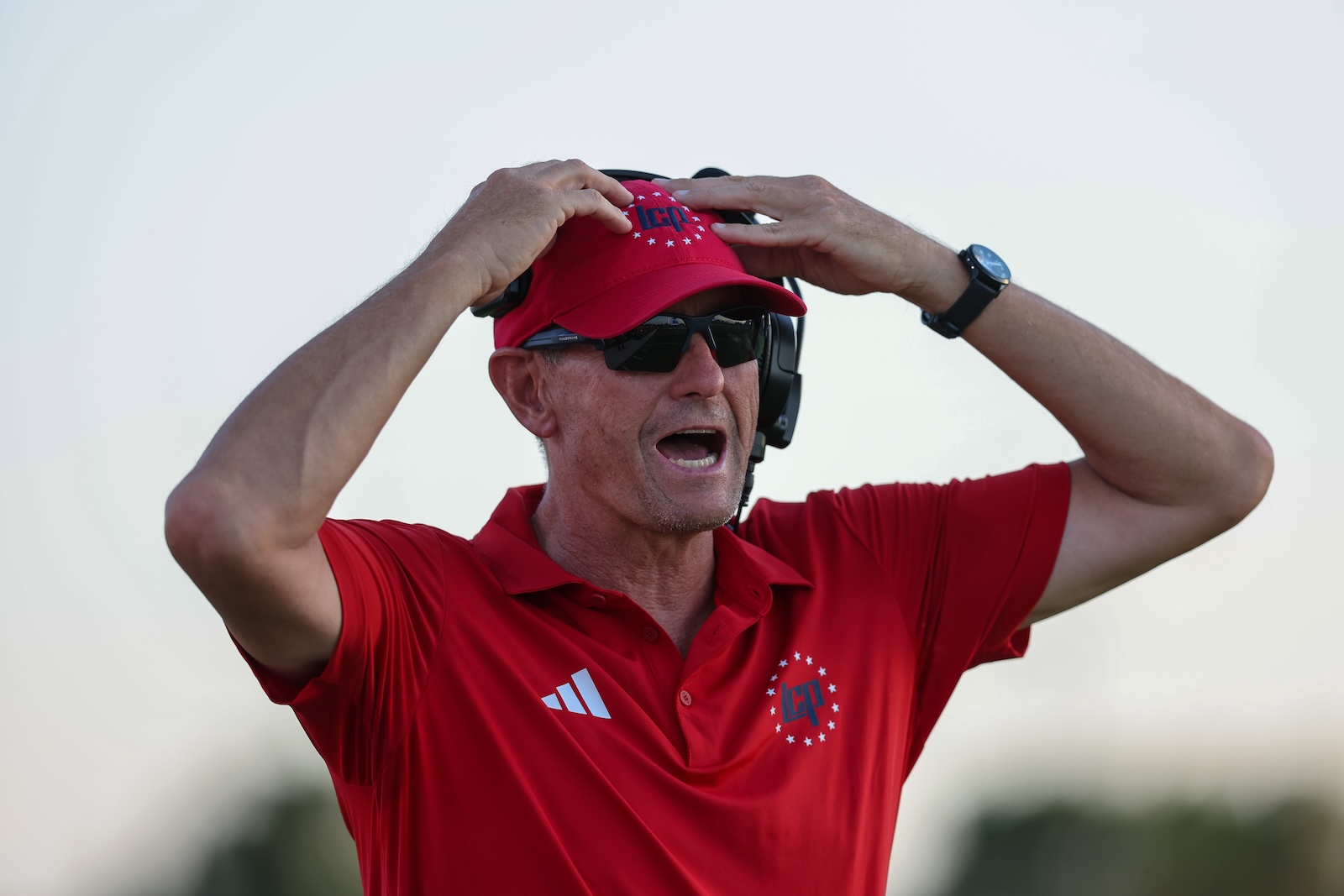Seabolt Chase-17282 | Press Pass Sports Coach in red polo shirt and cap, shouting with hands on his red cap, wearing sunglasses and a headset outdoors