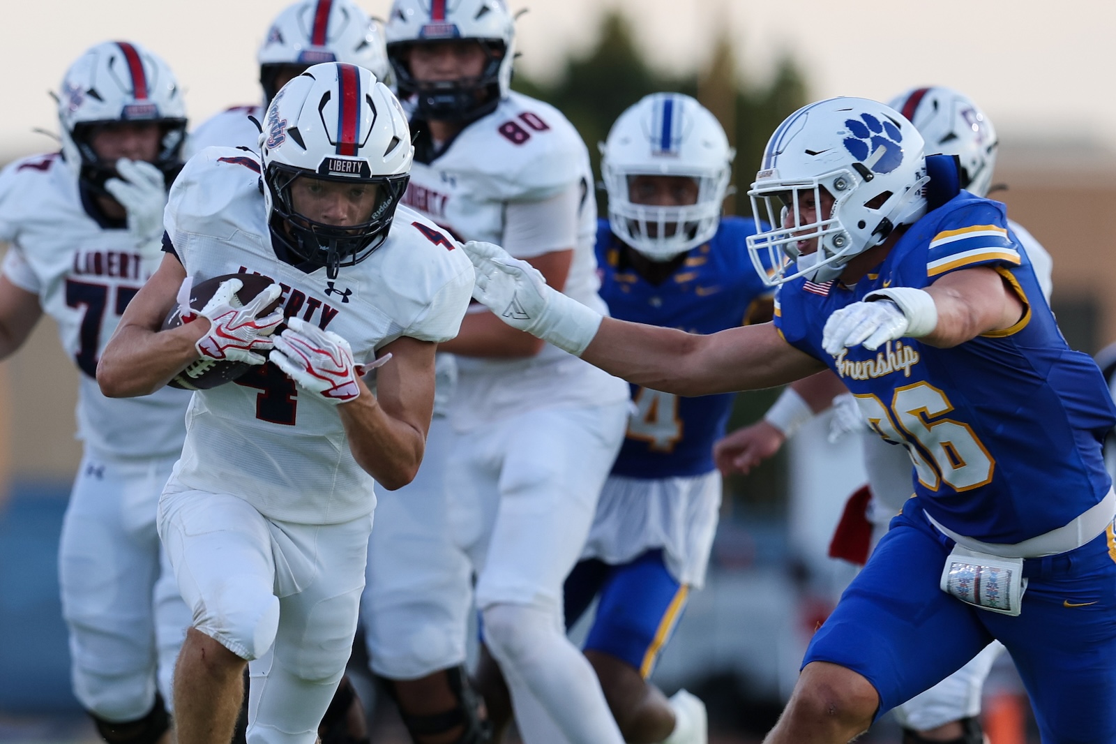 Seabolt Chase-17483 | Press Pass Sports African-American football player in a white Liberty uniform runs with the ball while a blue‑jersey defender reaches in for a tackle in pursuit behind him.