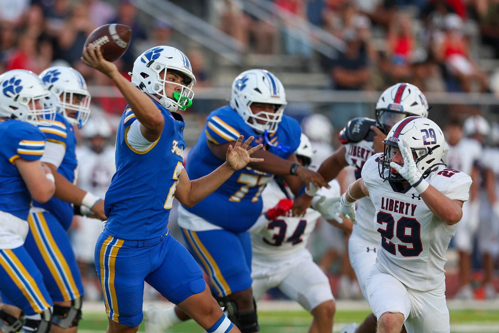 Seabolt Chase-17512 | Press Pass Sports A quarterback in a blue jersey throws a pass while defenders in white Liberty jerseys pursue him on a crowded field.
