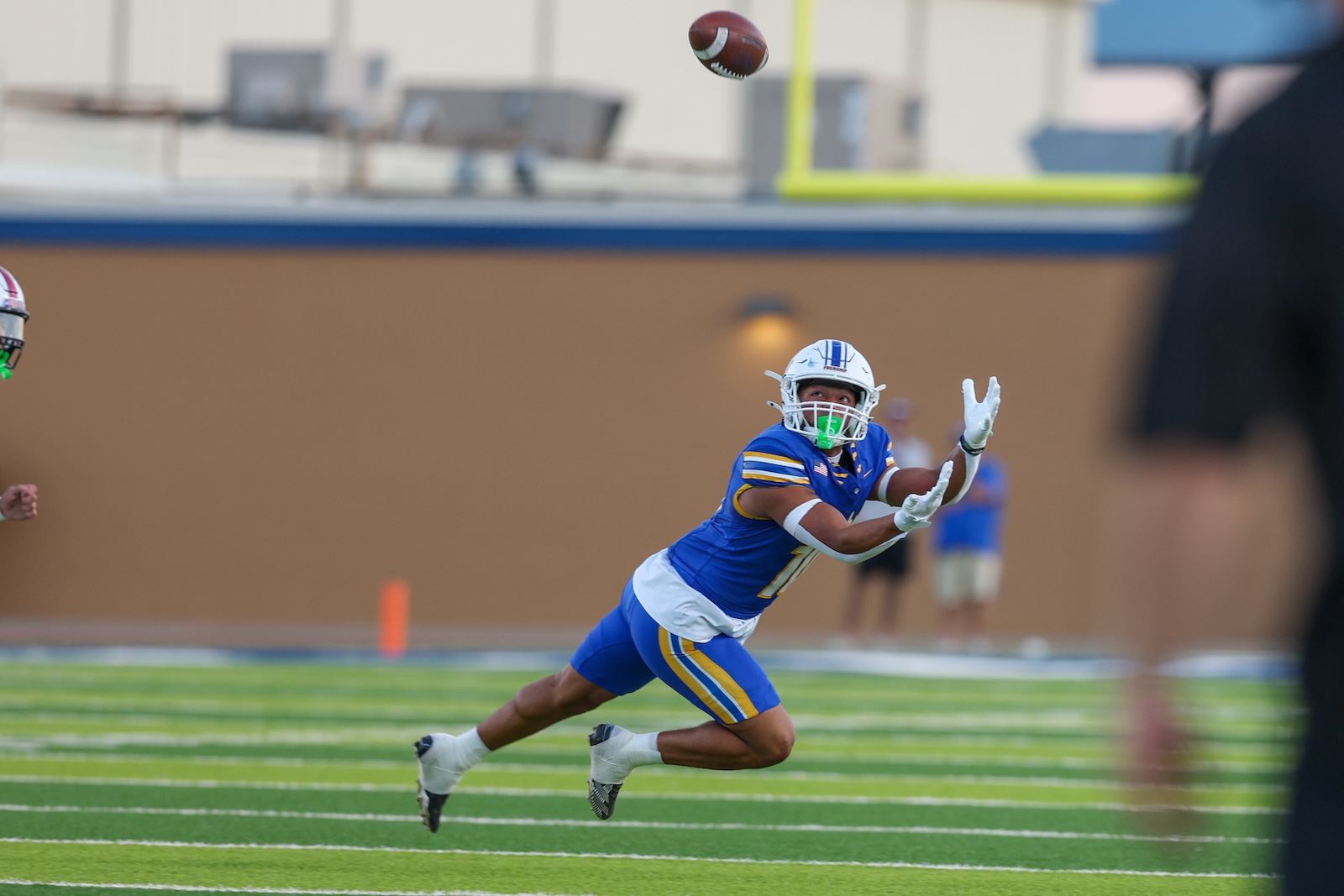 Seabolt Chase-17548 | Press Pass Sports Football player in blue and yellow dives to catch a spiraling ball on a green field.