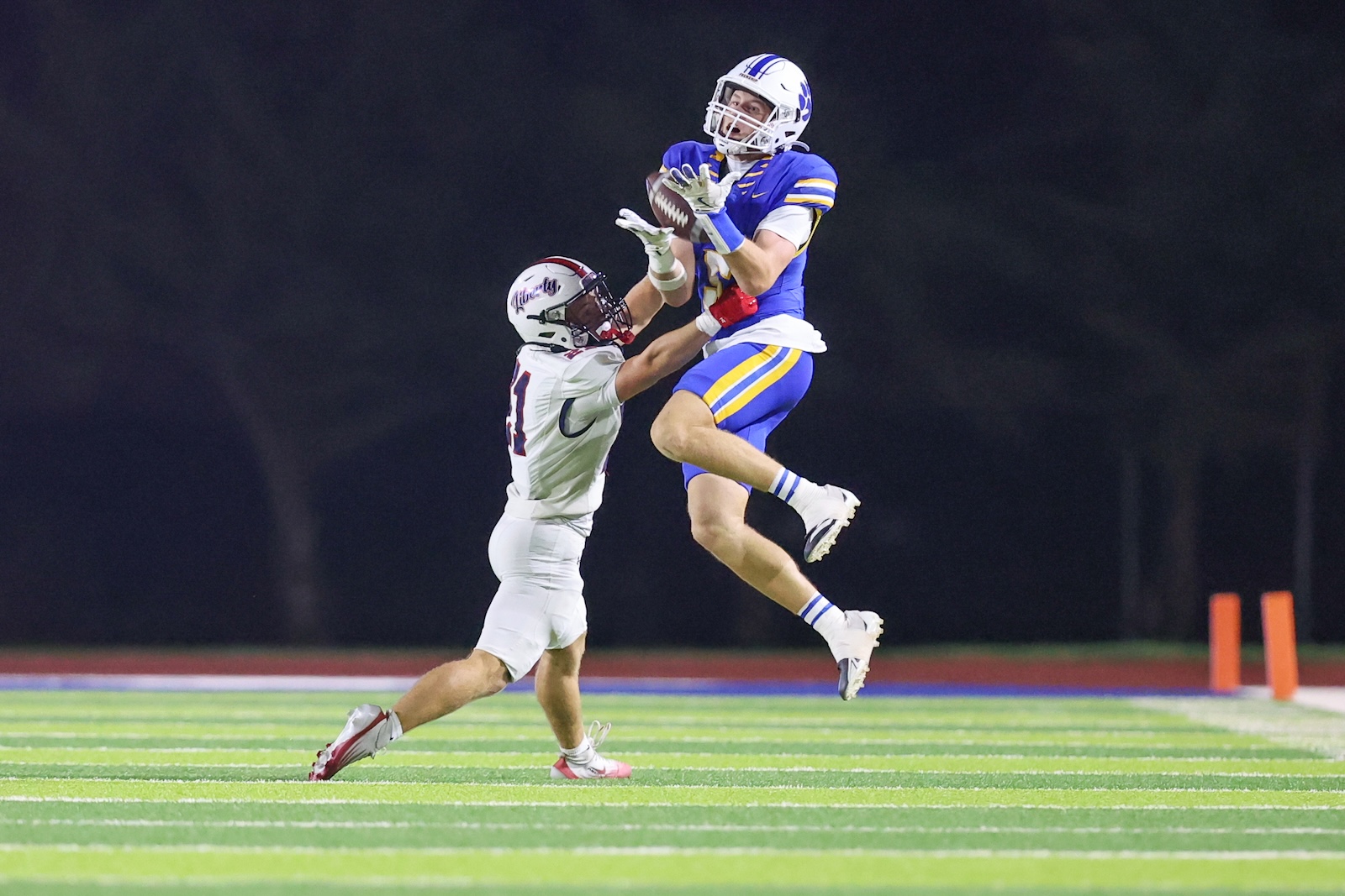 Number 5 frenship 21 liberty | Press Pass Sports Blue-uniform receiver leaps to catch a pass as a white-uniform defender reaches up during a night football game on a bright turf field.