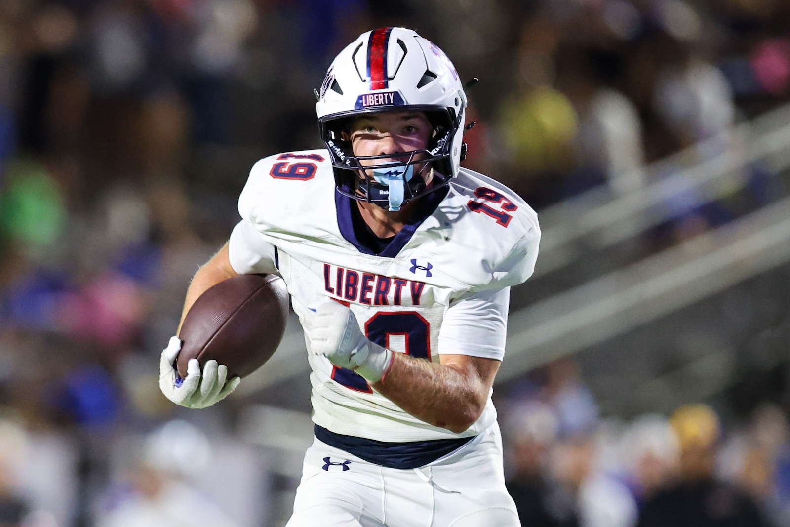 Seabolt Chase-18876 | Press Pass Sports Liberty football player in white jersey running with ball during a game, helmet on, crowd blurred in the background