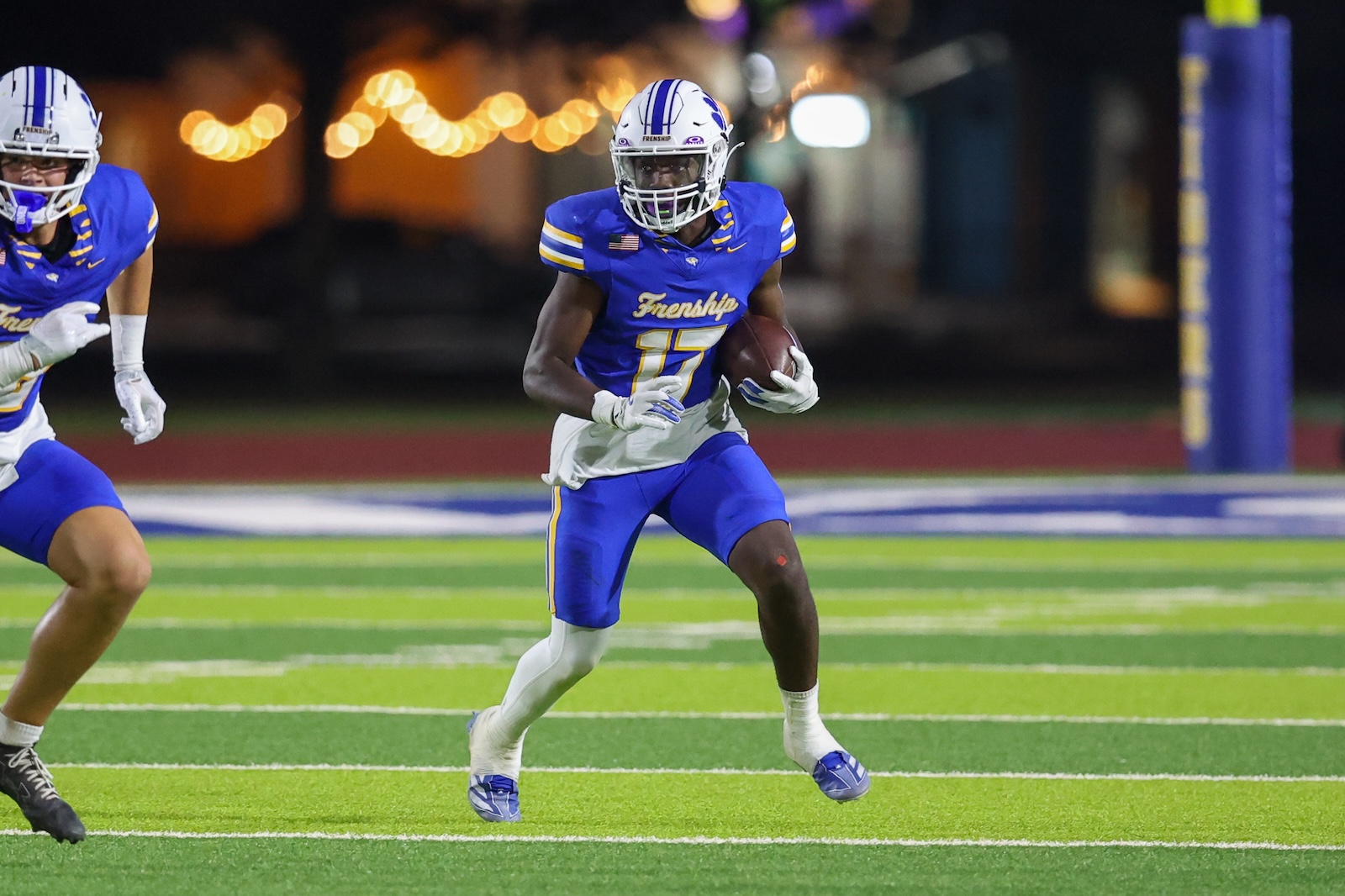 Seabolt Chase-18995 | Press Pass Sports Football player in blue and white uniform carrying the ball, number 15, running on a brightly lit grassy field at night with a teammate nearby.