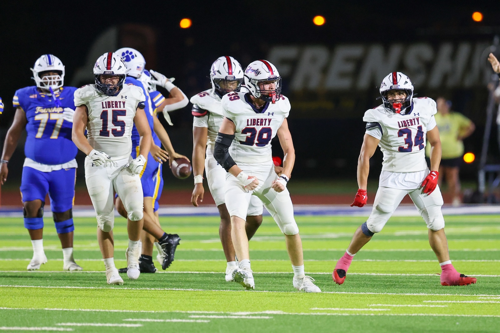 Seabolt Chase-19166 | Press Pass Sports Liberty football players in white jerseys sprint on a bright night field, with a blue-team opponent in the background.