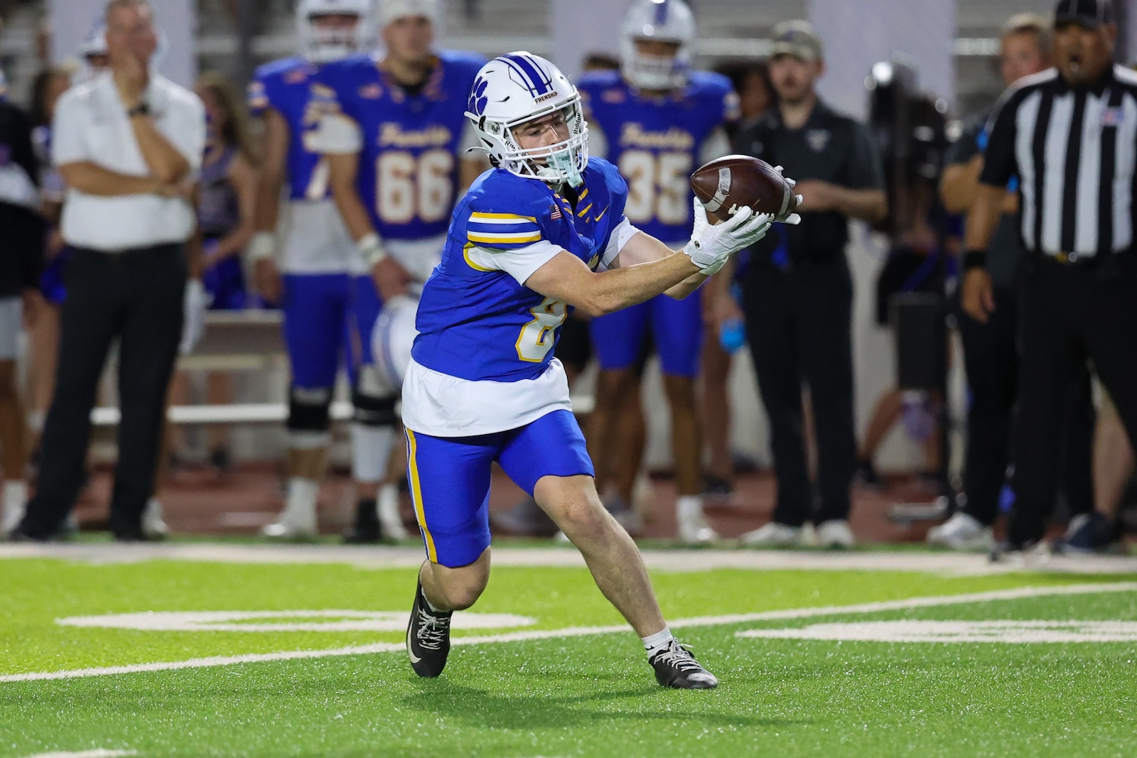 8 frenship | Press Pass Sports Football player in a blue and white uniform makes a catch on bright green turf with teammates and a referee in the background,