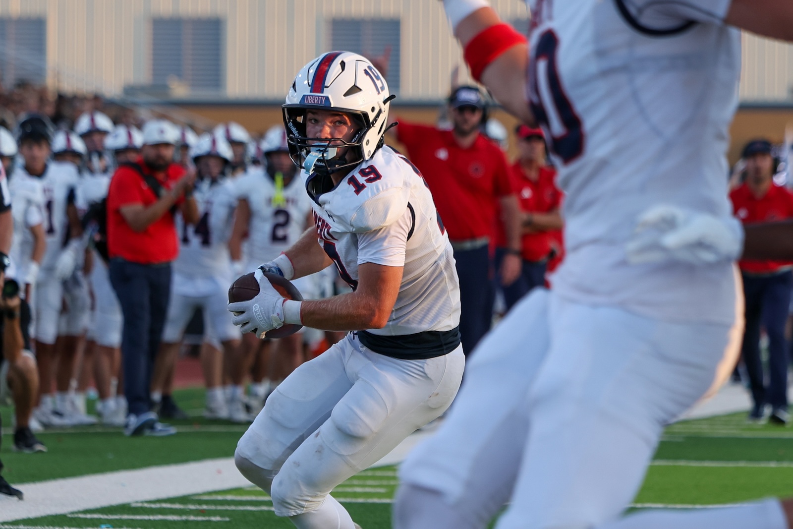Seabolt Chase-25417 | Press Pass Sports Football player in white jersey (number 19) runs with the ball during a game as teammates and fans watch from the sideline.