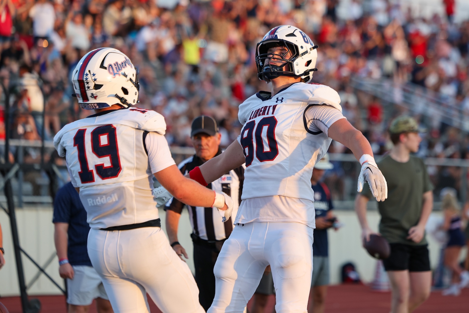 Seabolt Chase-25509 | Press Pass Sports Two football players in white Liberty uniforms (numbers 19 and 80) celebrate on the field as crowd cheers in the stands behind them.