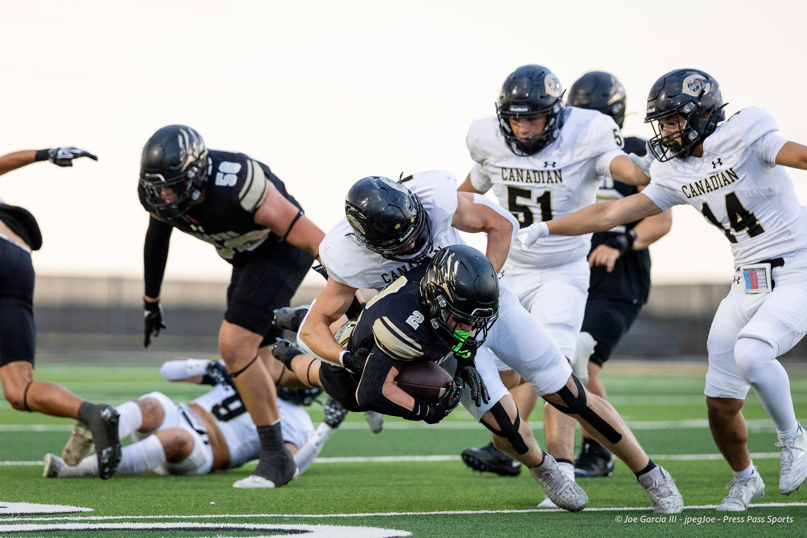 Canadian football players in white jerseys pursue a ball carrier in black as a tackle takes place on a green turf field.