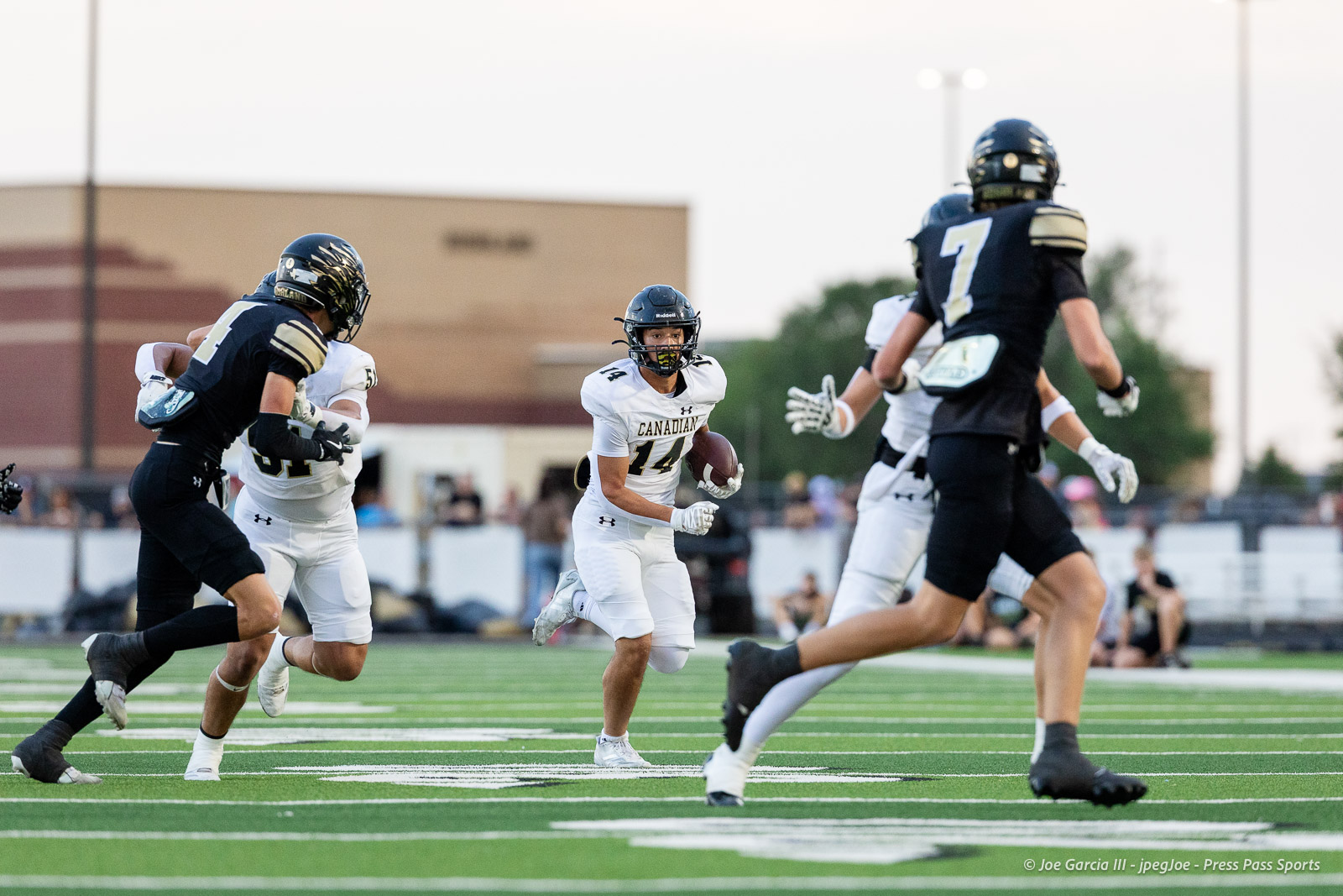 Canadian player #14 in white carries the ball as defenders in black chase him on a green football field during a game moment.