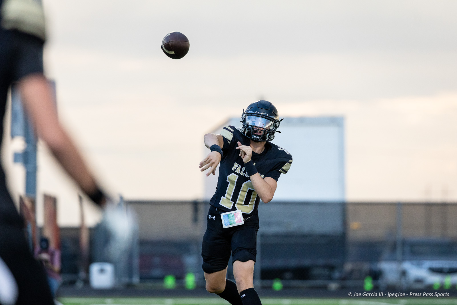 Quarterback in black and gold with number 10 mid-throw during a football game on a sunny day.
