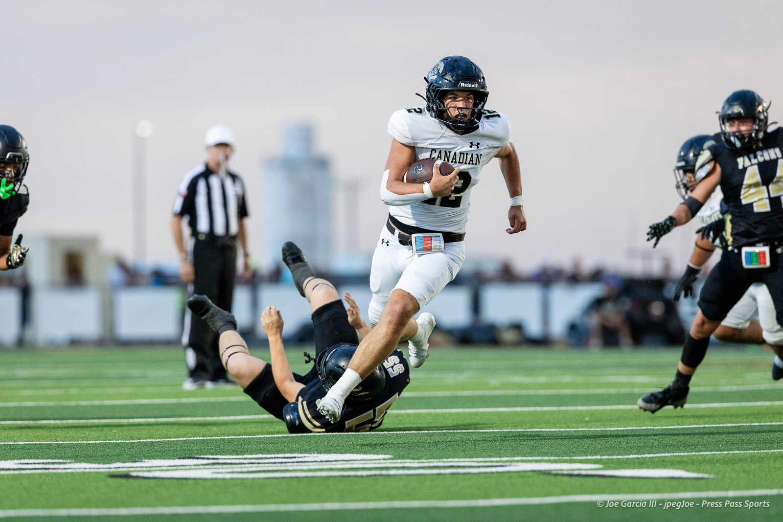 White-uniform football player (#2) carries the ball as an opposing defender tumbles on the artificial turf behind him.