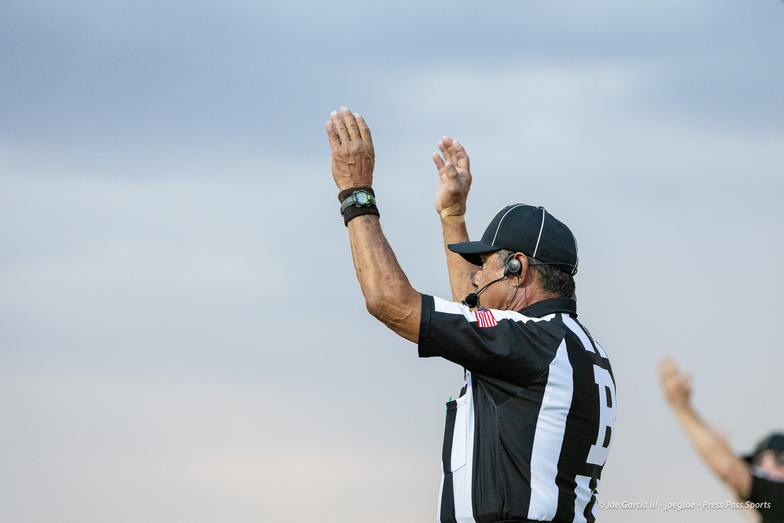 American football referee raises both arms to signal a touchdown against a cloudy sky backdrop.